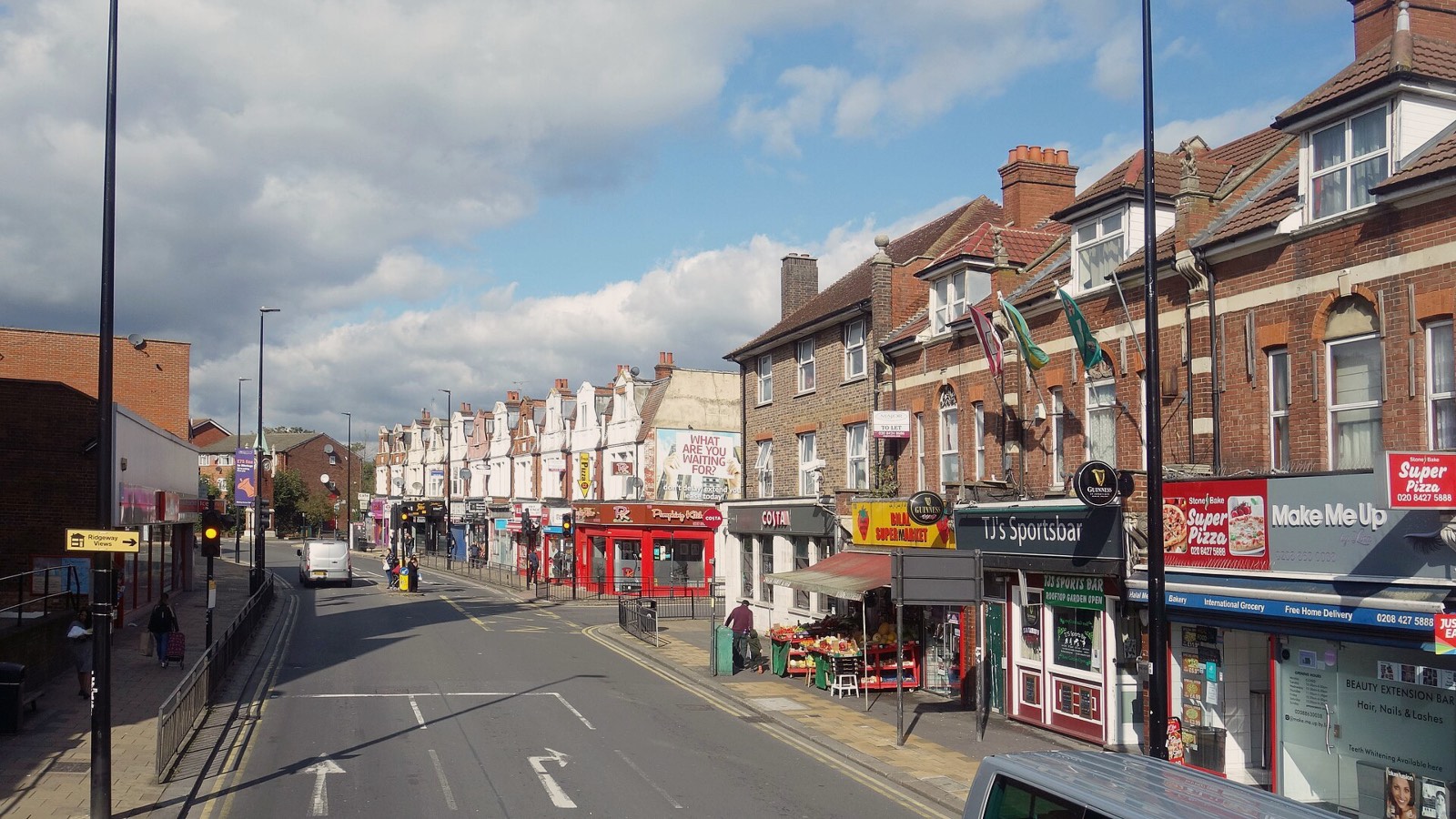 A British high street seen from a bus top deck: rows of small shops, a greengrocer's stall and parked vans under a cloudy blue sky.