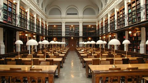 A long library reading room with tables and green lamps