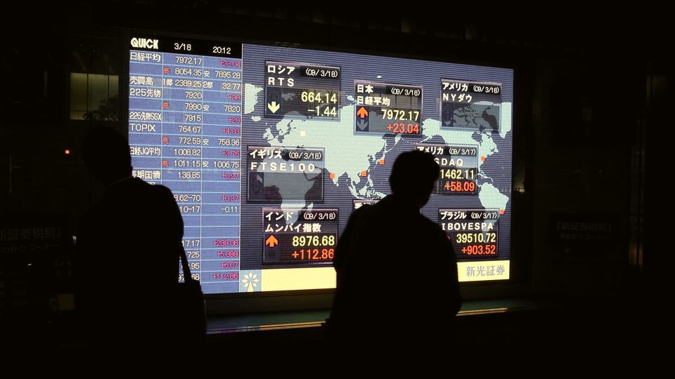 Two people in silhouette watching a large electronic board of share prices at night